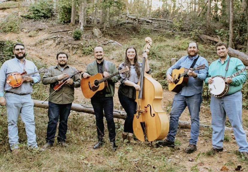 The 6 members of the 700 South bluegrass band standing in the woods with their instruments. 